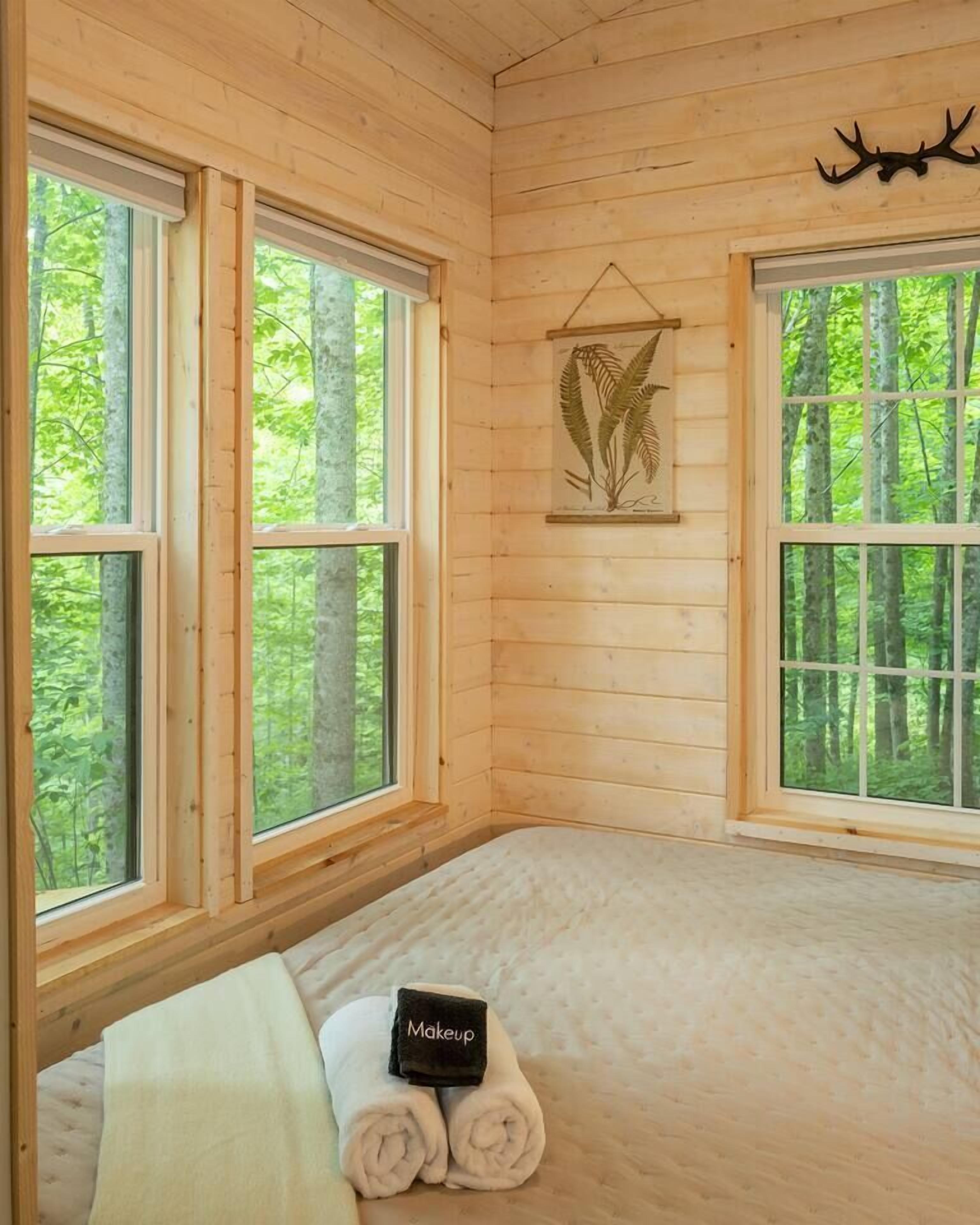 Une pièce de style cabane en bois, dotée de grandes fenêtres, d’un lit, d’une décoration murale représentant une tête de cerf et d’une œuvre d’art représentant une fougère.