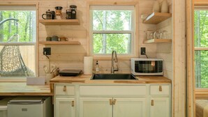 A compact kitchen with wooden cabinets, a microwave, a sink, and various kitchen utensils on shelves. - Serene, Romantic Getaway, Outdoor Shower, and Hiking! (Sylva)