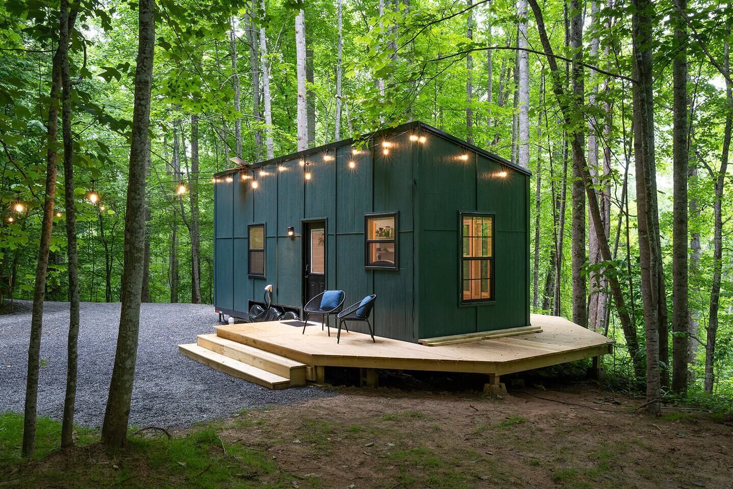 Una cabaña pequeña de color verde oscuro con una terraza de madera, dos sillas y luces colgantes.