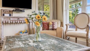 Ein Wohnzimmer mit einem rustikalen Holztisch, einer Couch mit gemusterten Kissen, einem Holzstuhl, einem an der Wand befestigten Fernseher und einer Vase mit Sonnenblumen.