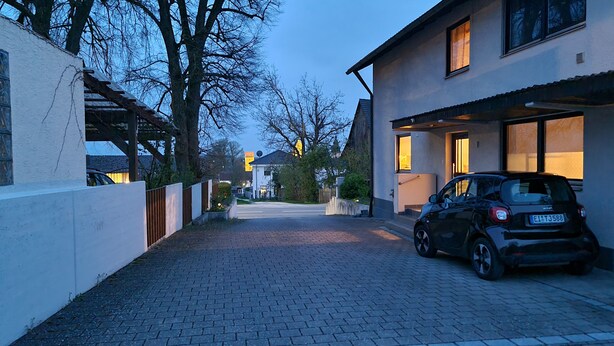 Una calle residencial con entrada pavimentada, un coche aparcado y casas con ventanas iluminadas.