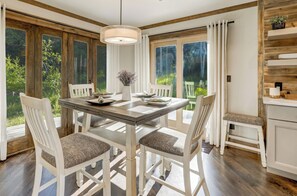Un comedor con mesa de madera, sillas blancas y vistas a la vegetación a través de amplias ventanas.