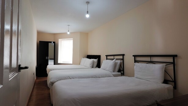 A hotel room with three single beds, a wooden floor, a window with blinds, and a door leading to another room. - The Serenity Apartment (Manchester)