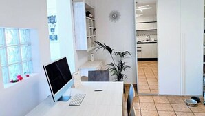 Een moderne keuken met witte kasten, een eethoek met een witte tafel en stoelen, en een bureau met een monitor en toetsenbord.