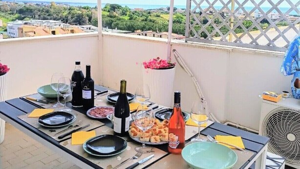 Een balkon met een tafel gedekt voor twee personen, met wijnflessen, borden en een ventilator.