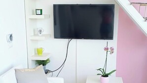 Een woonkamer met een witte bank, een klein wit tafeltje, een roze bloempot en een tv aan de muur.