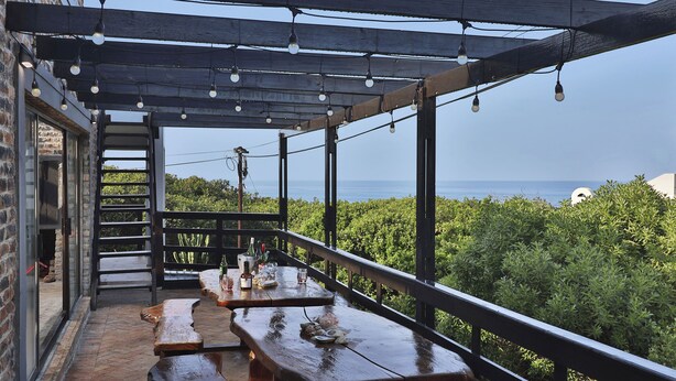 Un patio al aire libre con mesas y bancos de madera, una pérgola con luces colgantes y vistas al mar.