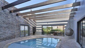 Una piscina con borde de ladrillo, una pérgola de madera sobre ella y un muro de piedra.