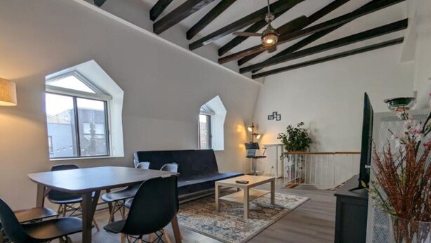 A modern living room with a dining area, featuring a large table, chairs, a sofa, and a coffee table. There are windows providing natural light and a view of the outside.
