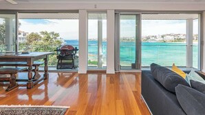 A spacious living area with a wooden floor, a dark sofa, and a view of the sea through large glass doors. - Ocean view luxury 3 bedroom home with balcony (North Bondi)