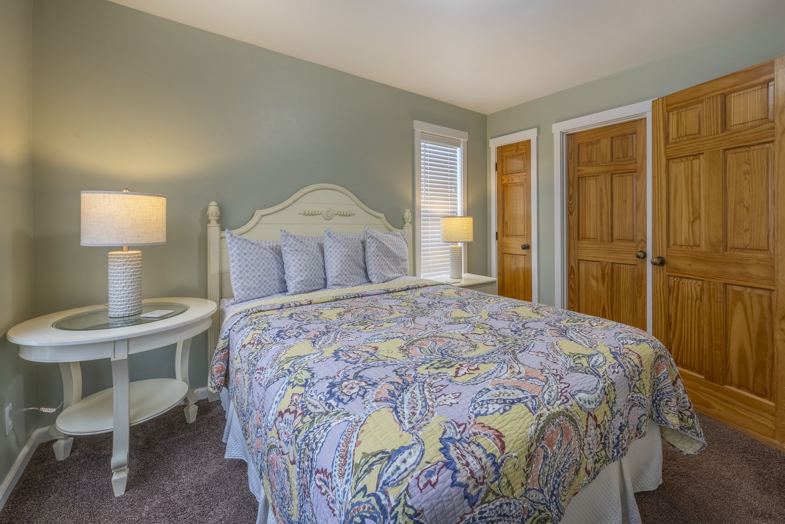 Une chambre à coucher comprenant un lit, deux tables de chevet, une lampe, une fenêtre avec des stores et une porte en bois.