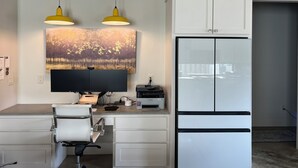 A modern kitchen with a white refrigerator, a flat-screen TV, a dining table with chairs, and two pendant lights.