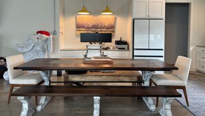 A modern kitchen with a wooden dining table, two chairs, and a bench. There is a large flat-screen TV mounted above the counter, a refrigerator, and a painting on the wall.
