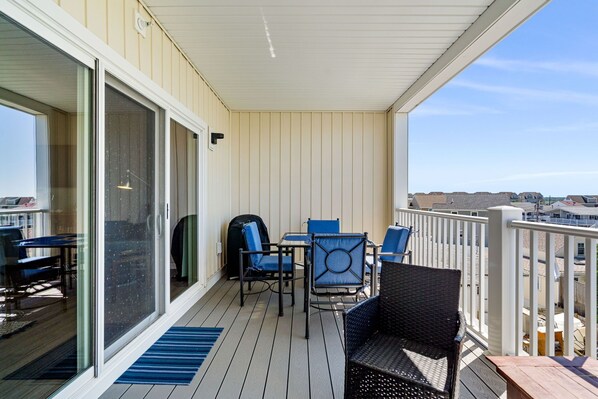 A covered outdoor area with a table and chairs, a wicker chair, and a view of the beach and other buildings.