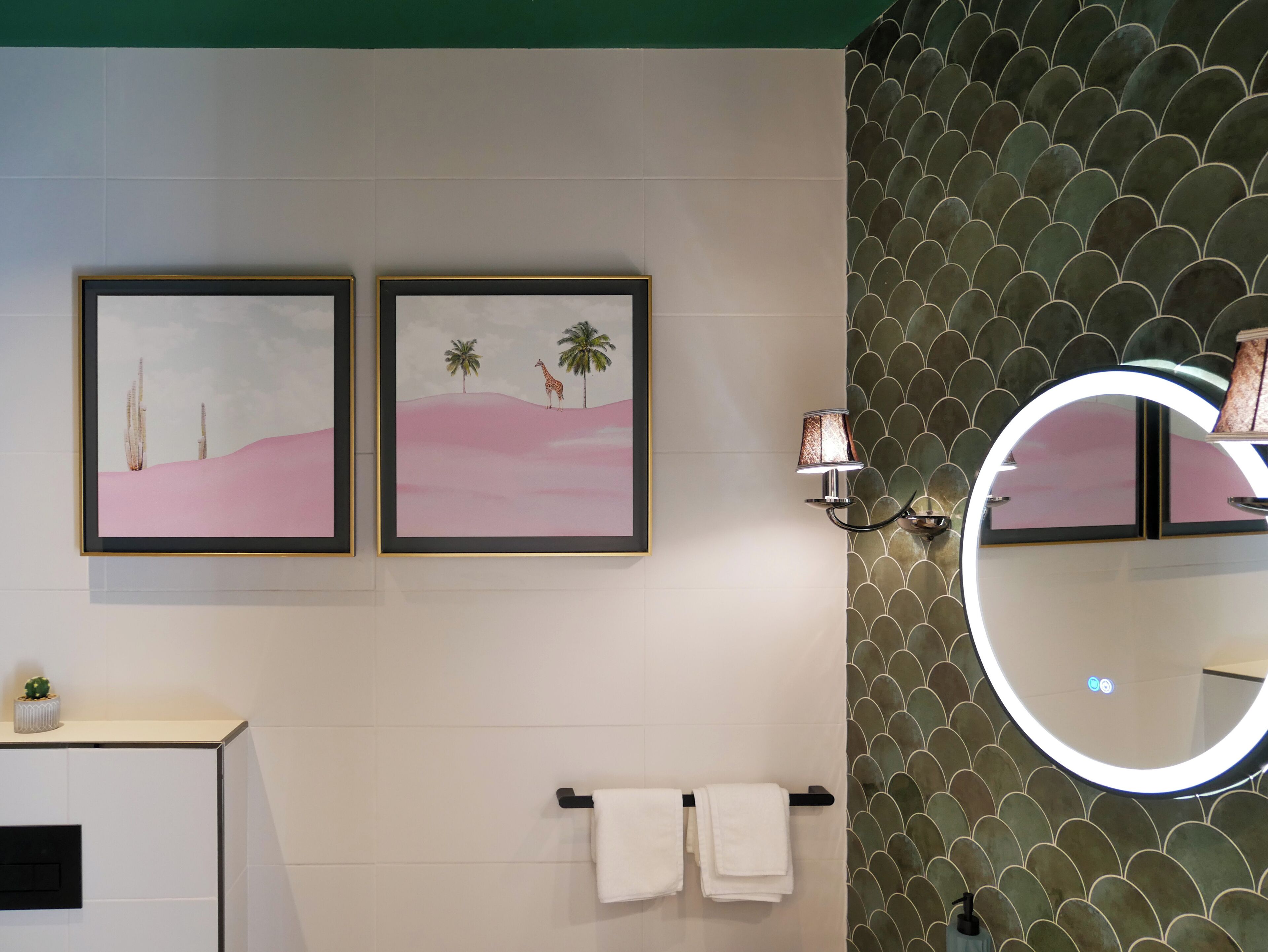 Une salle de bain avec deux tableaux encadrés, un porte-serviettes avec des serviettes blanches, un miroir avec un éclairage circulaire et un mur à motifs verts.