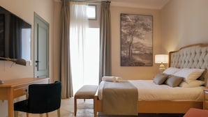 Una camera d'albergo con un letto, una scrivania, una sedia, una televisione, una finestra con tende e un quadro appeso al muro.