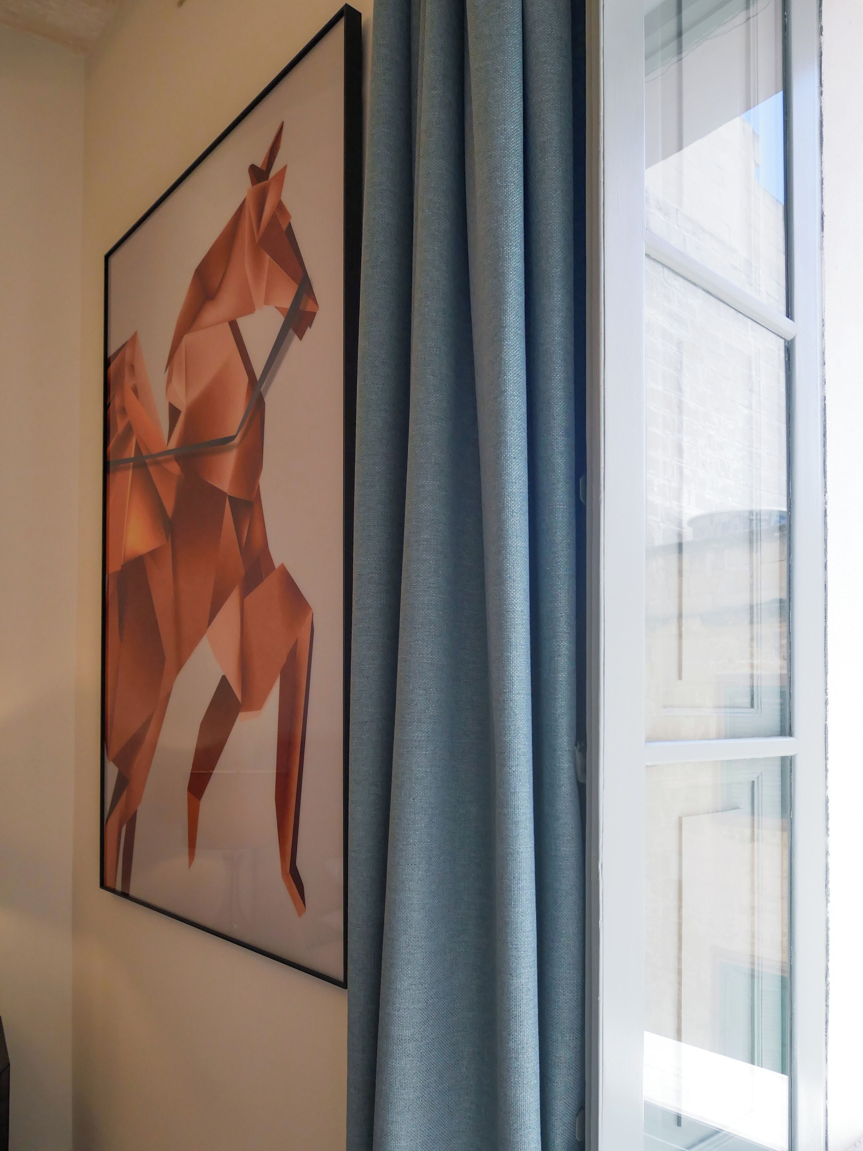 Uma pintura de um cavalo geométrico emoldurada numa parede, uma janela com persianas e uma cortina azul.
