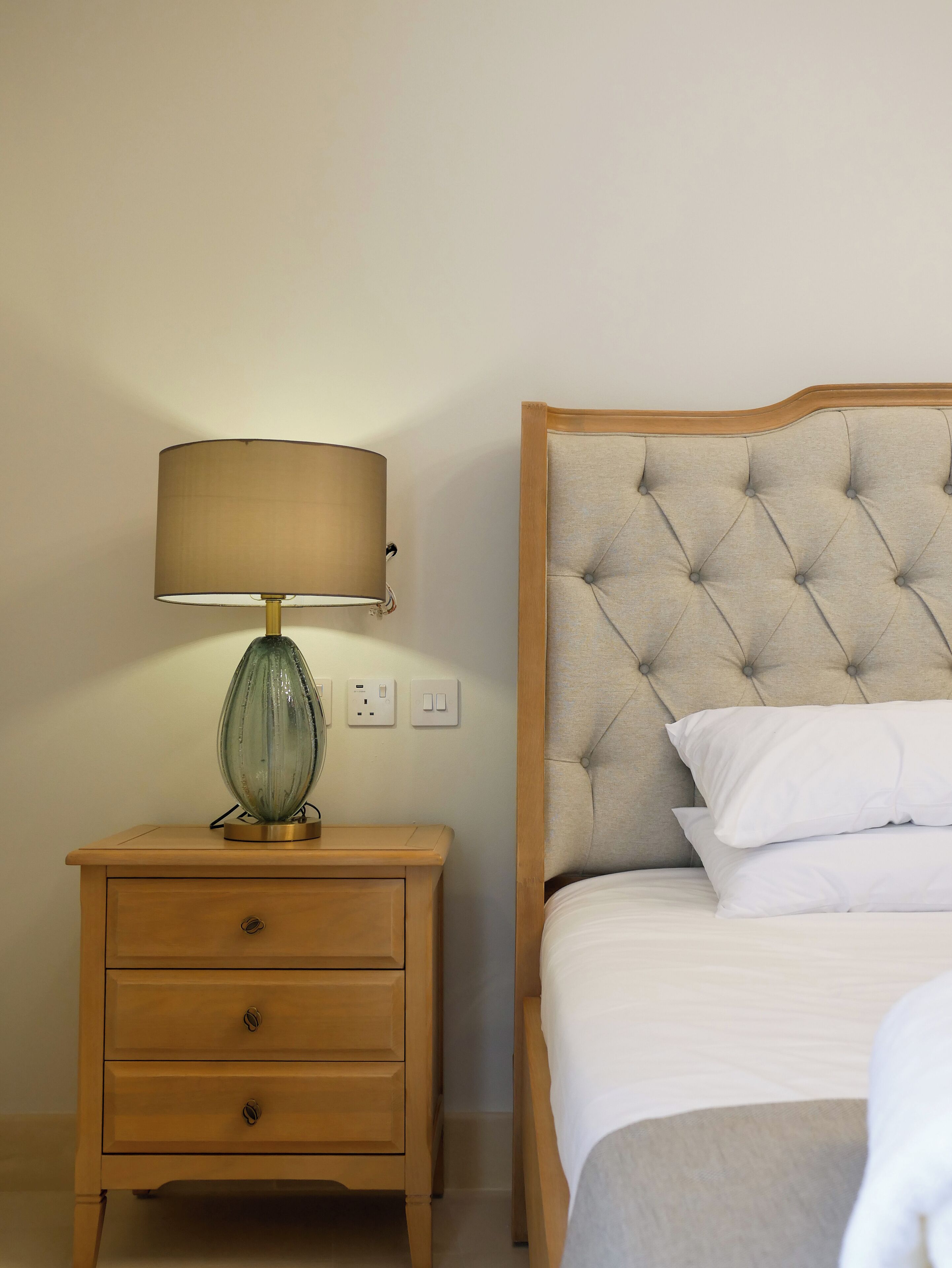 Una cama bien tendida con sábanas blancas y un cabecero capitonado, una mesita de noche con una lámpara y un objeto de vidrio decorativo, y un mueble auxiliar de madera con cajones.