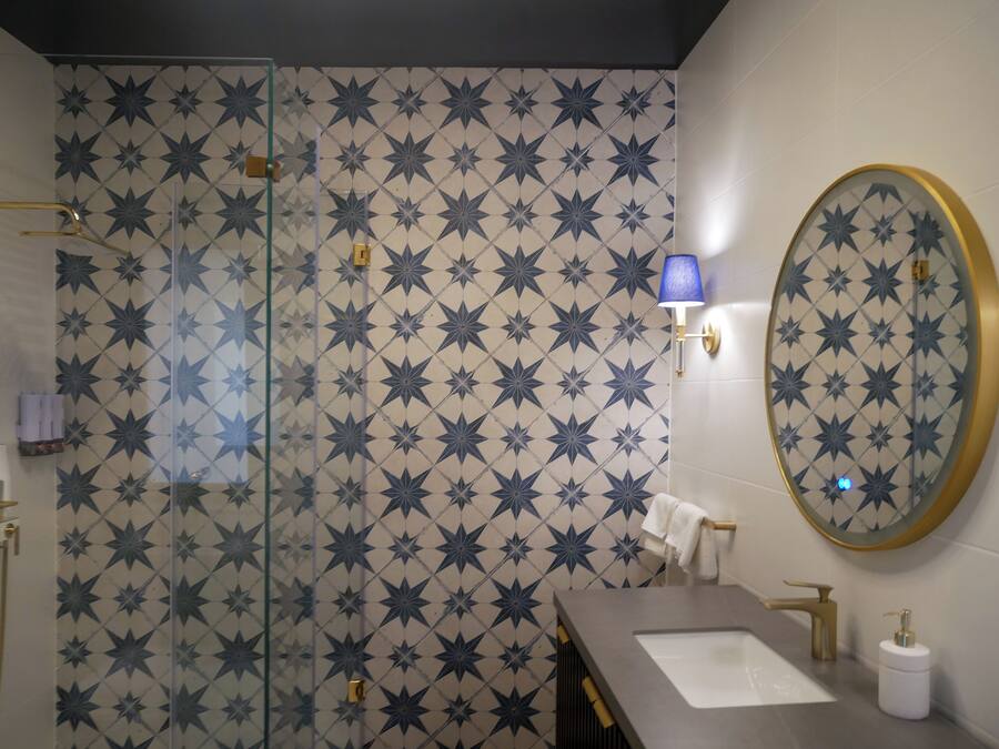 Un baño con azulejos estampados con motivos de estrellas, cerramiento de ducha de cristal, lavabo con grifo dorado, lámpara azul en la pared y espejo redondo con marco dorado.