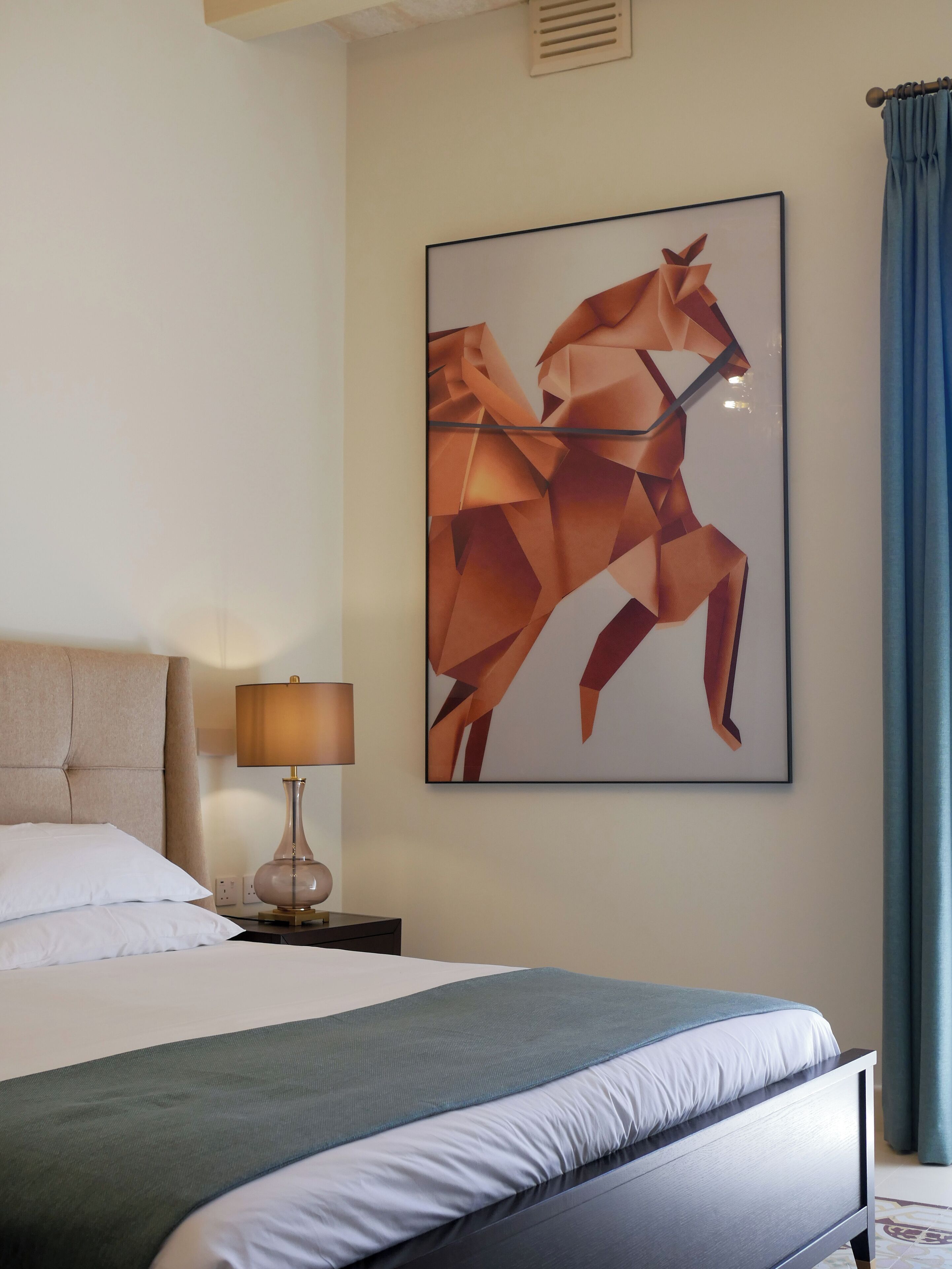 Una cama bien hecha con un cubrecolcha verde, una lámpara de noche y un cuadro abstracto enmarcado en la pared.