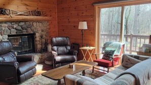 L’intérieur chaleureux d’un chalet avec une cheminée en pierre, des fauteuils en cuir et une grande fenêtre donnant sur une forêt.