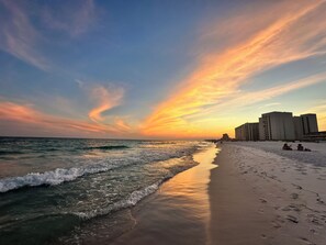 A beach with a sunset, ocean waves, and buildings in the background.