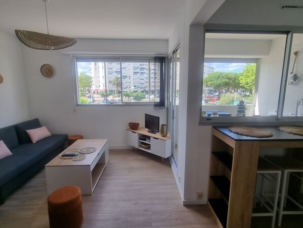 Een moderne woonkamer met een bank, salontafel, televisie en een groot raam met uitzicht op gebouwen en bomen.