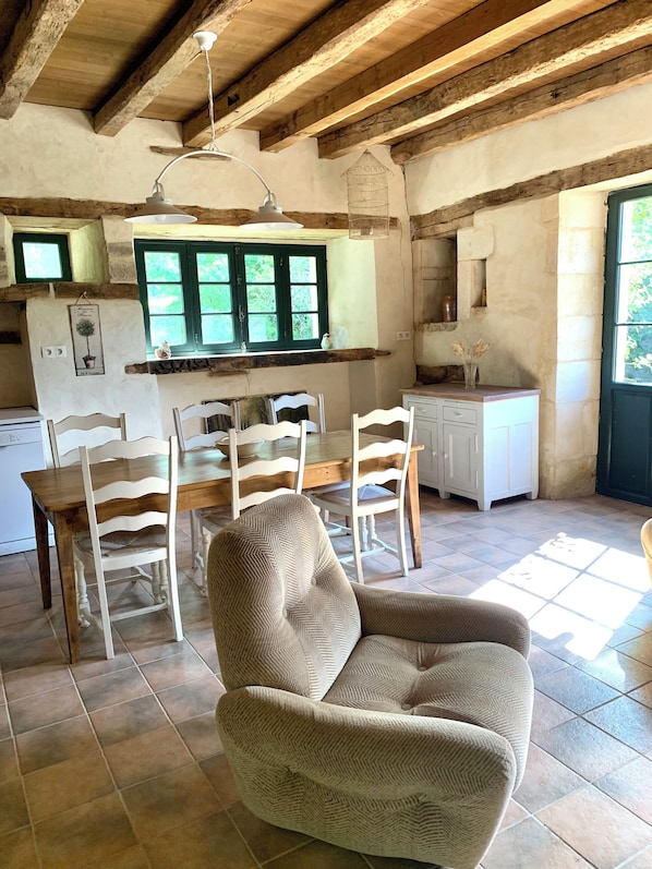 A rustic dining area with wooden chairs and a table, a kitchen with a window, and a hanging light fixture. - Gite Simple et Authentique, au Calme Pour les Amoureux de la Nature (Bazoges-en-Pareds)