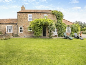 A brick house with a tiled roof, a front door, and a small garden with a lawn and two chairs.