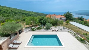 A rectangular swimming pool with lounge chairs, surrounded by greenery and a view of a coastal landscape. - Apartment Klara - beautiful pool apartment for 7 people (Rabac)