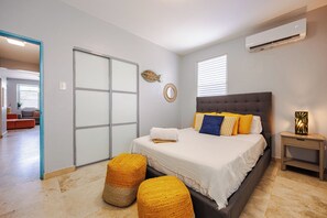 Ein Schlafzimmer mit einem Bett, einem Nachttisch, einer Wand-Klimaanlage und Blick auf den angrenzenden Wohnbereich.