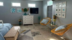 Ein Schlafzimmer mit einem Bett, zwei Korbstühlen, einem Schminktisch und einem Fernseher. An den Wänden befinden sich Kunstwerke.