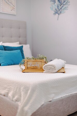 Una cama bien hecha con edredón blanco, una caja de madera con la palabra 'Family' y una toalla blanca enrollada sobre un taburete de madera.