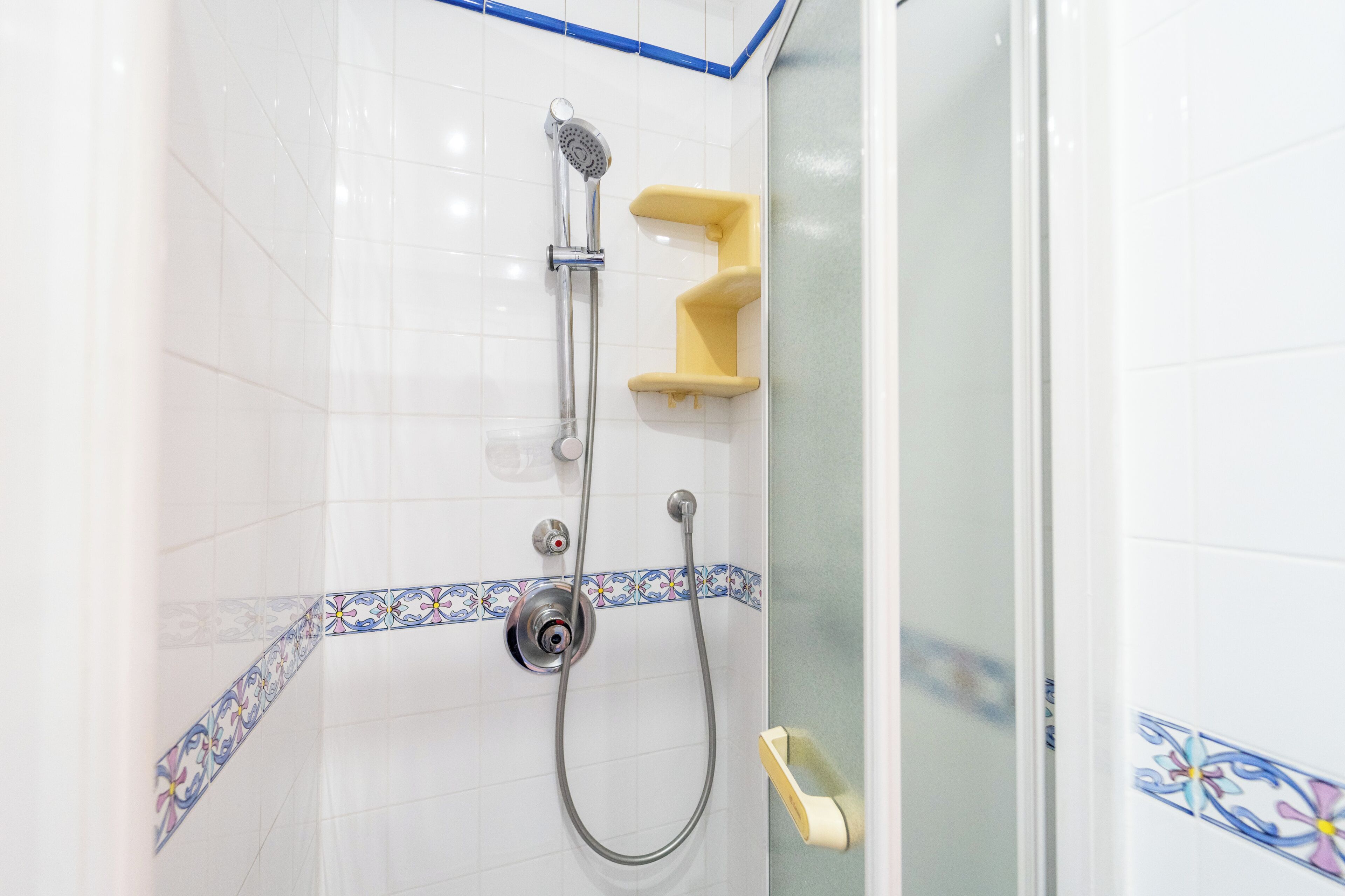 Ducha con cabezal de ducha de mano y grifo empotrado, un estante en la esquina y un borde decorativo de azulejos.