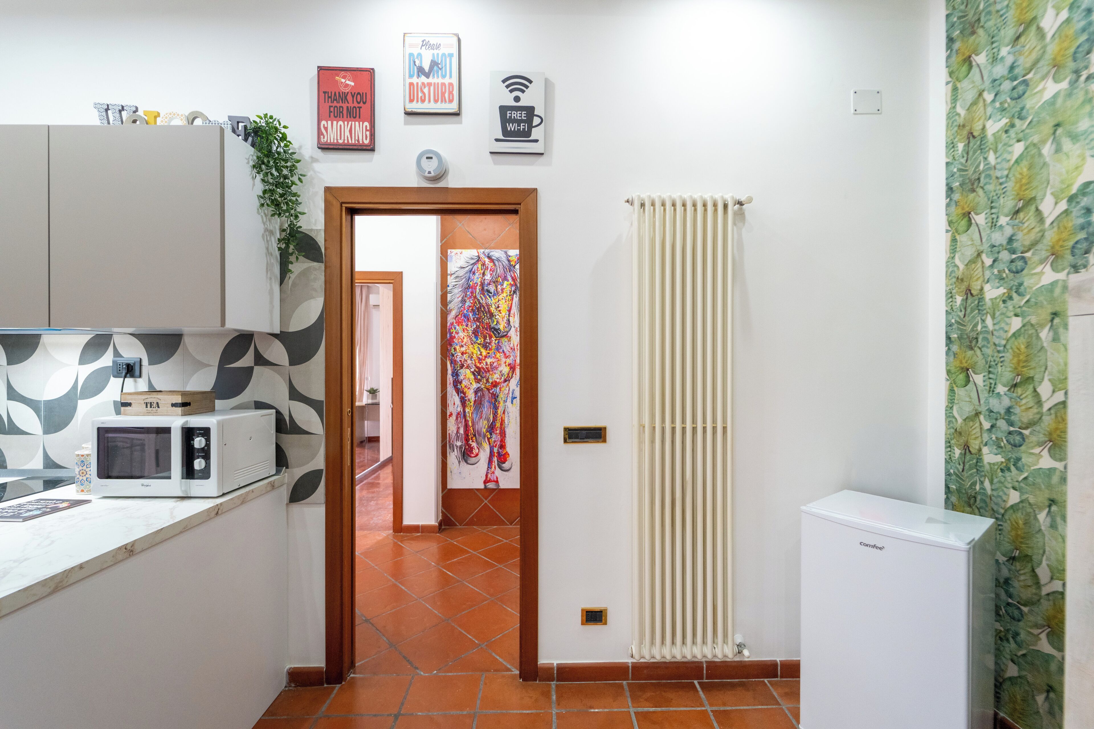 A compact kitchen with a microwave, a small refrigerator, and a door leading to another room. There are wall-mounted signs, a plant, and a radiator.