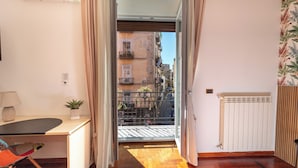 Une pièce avec un plancher en bois, un bureau avec une lampe, une chaise, une plante en pot et une vue sur un balcon doté d’une balustrade, avec un immeuble en arrière-plan.
