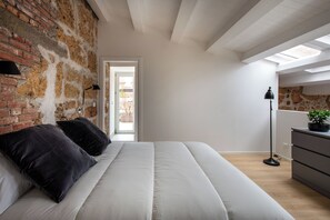 Camera da letto con parete in mattoni, lucernario, letto con biancheria bianca, lampada da terra e cassettiera.