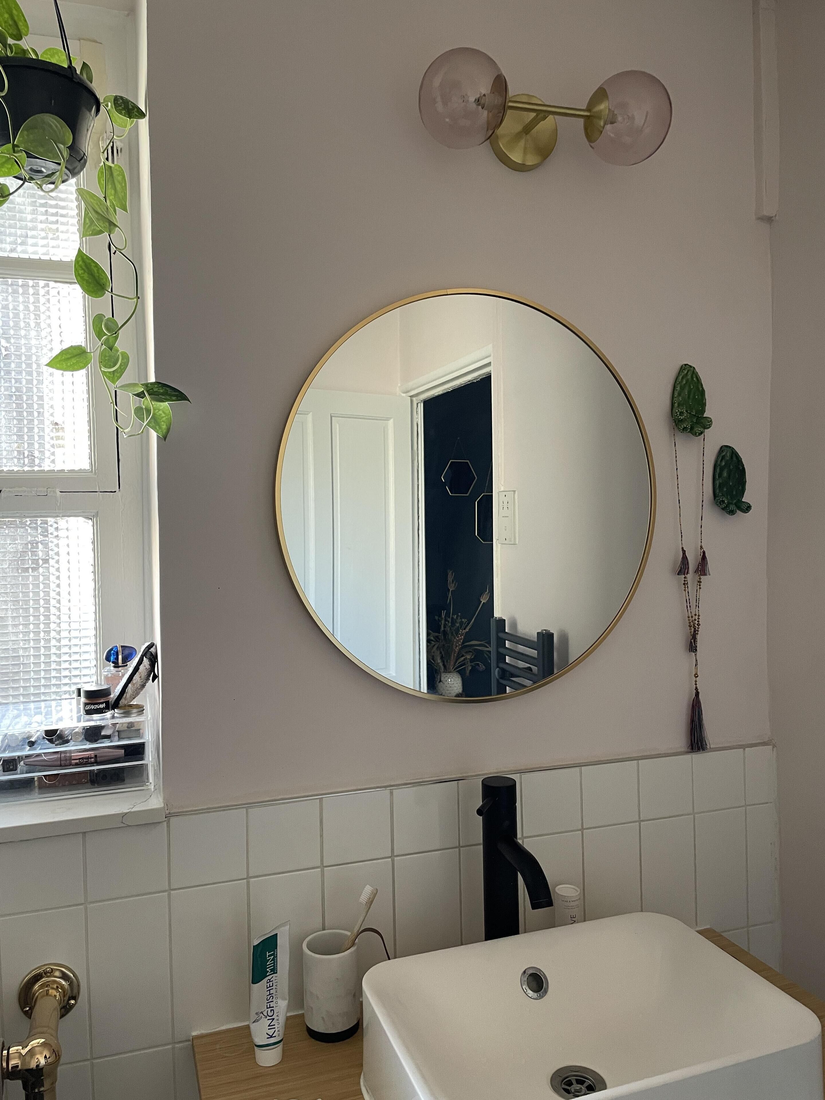 A bathroom with a white sink, a black faucet, a round mirror, and a hanging plant.