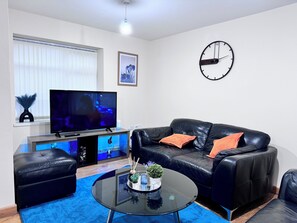 Una sala de estar moderna con un sofá de cuero negro, una mesa de centro de vidrio, un televisor de pantalla plana y un reloj de pared.