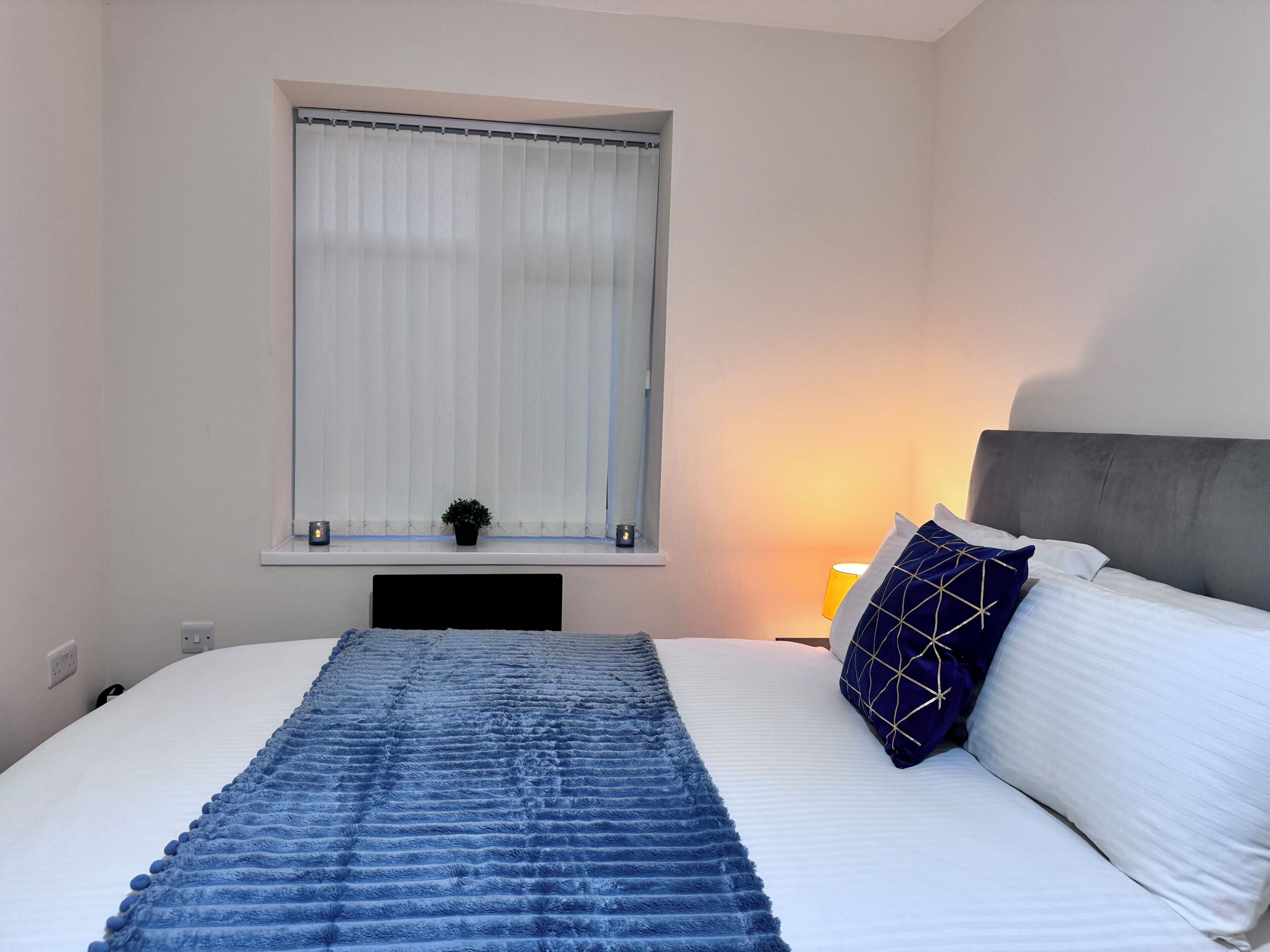 A bedroom with a bed, a window with blinds, a bedside lamp, and a small plant on the windowsill.