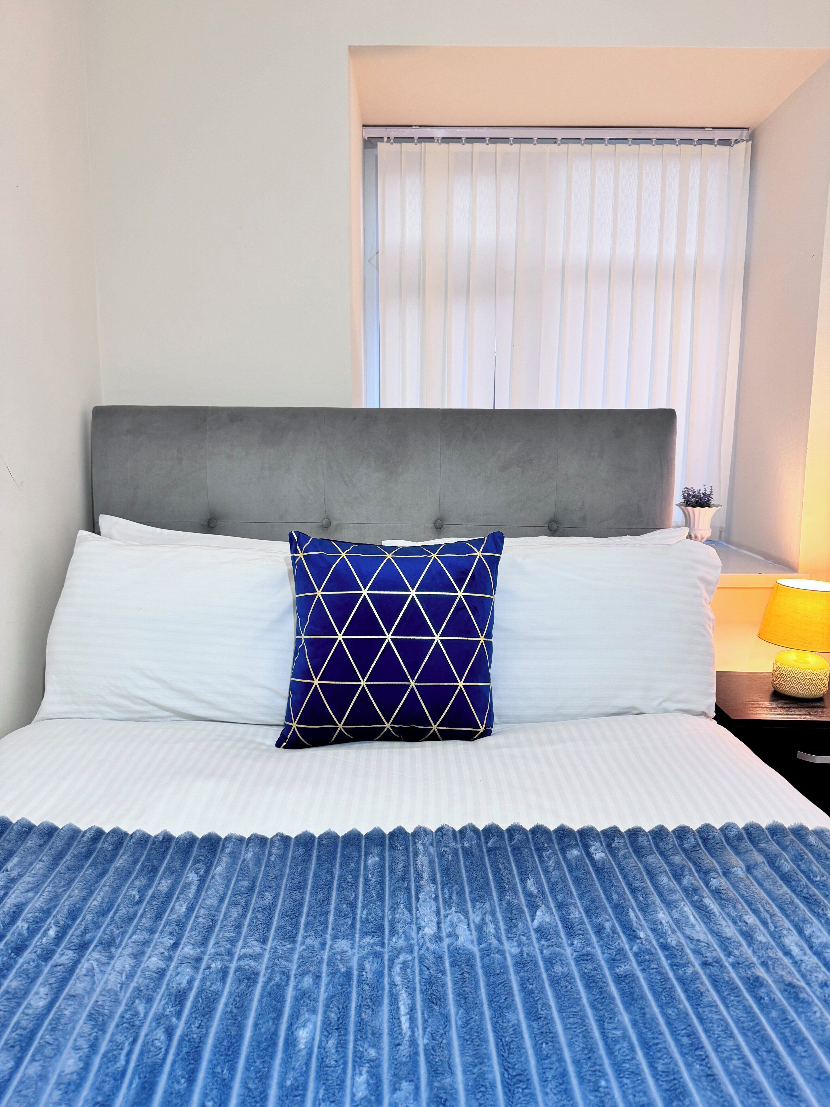 A neatly made bed with a grey headboard, white linens, and a blue patterned pillow.