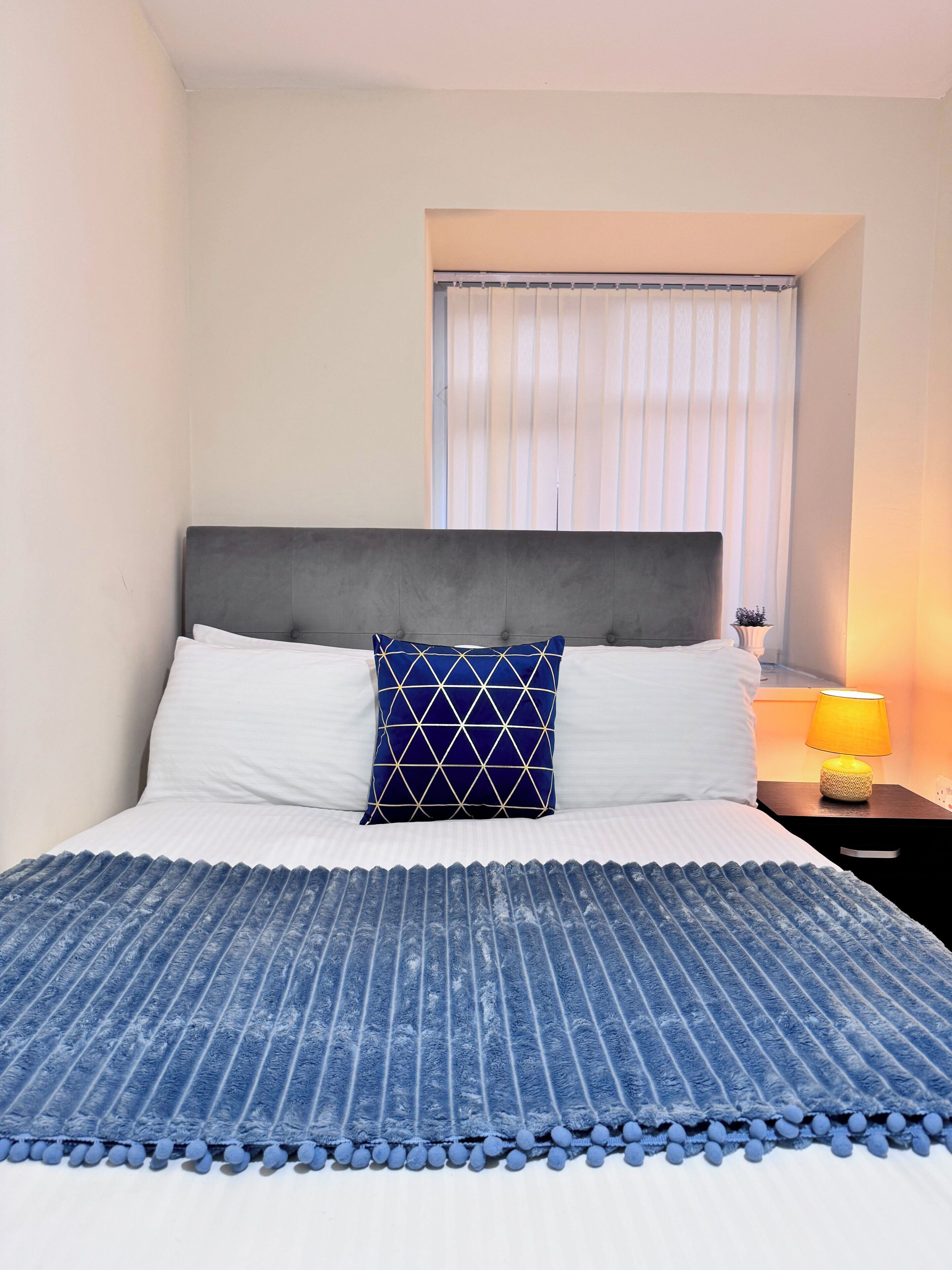 Un lit bien fait, recouvert d’une couvre-lit à motifs bleus et blancs, avec une tête de lit rembourrée de couleur grise et un seul coussin décoratif à motifs géométriques.