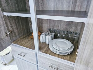 Un mueble de madera con estantes que contienen botellas de vidrio, una taza blanca y platos apilados.