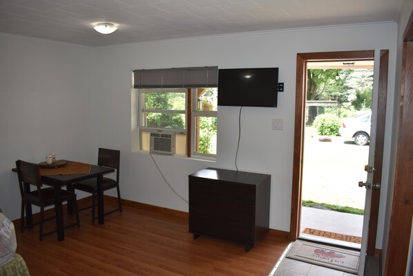 A room with a wooden floor, a dining table with chairs, a black dresser, a TV mounted on the wall, and a door leading outside. - PL Motel (Greentown)