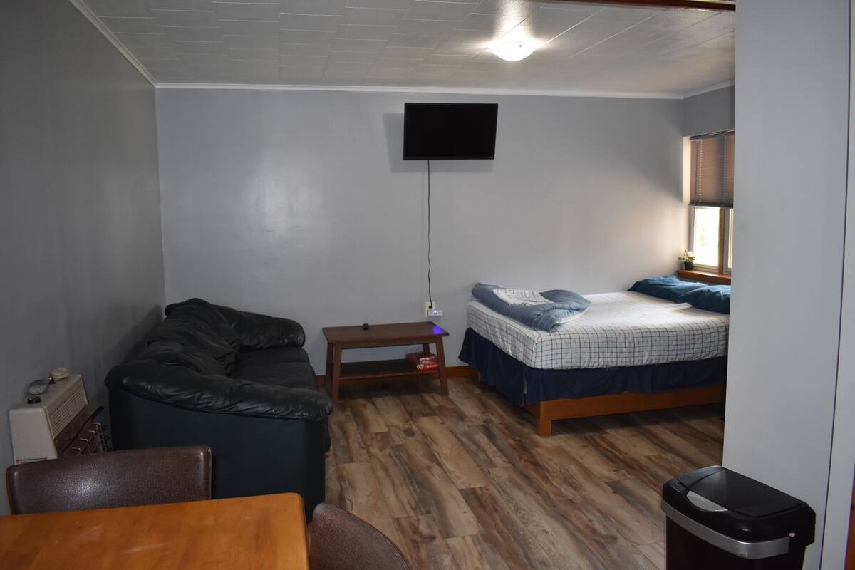 Una habitación pequeña y funcional con una cama individual, un sofá, una mesa y un televisor montado en la pared.