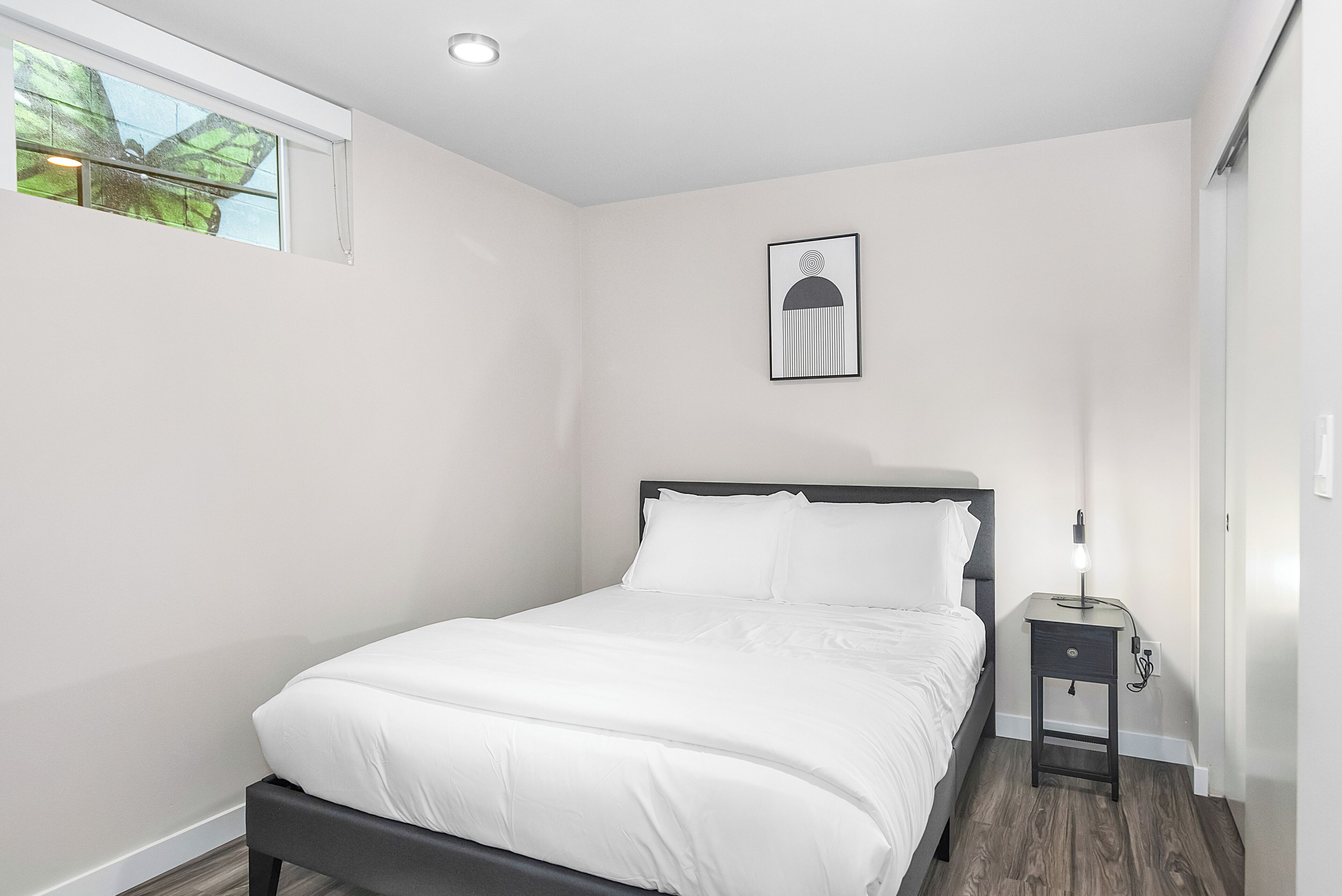 A bedroom with a bed, a nightstand, a wall-mounted light, and a window with a view of trees.