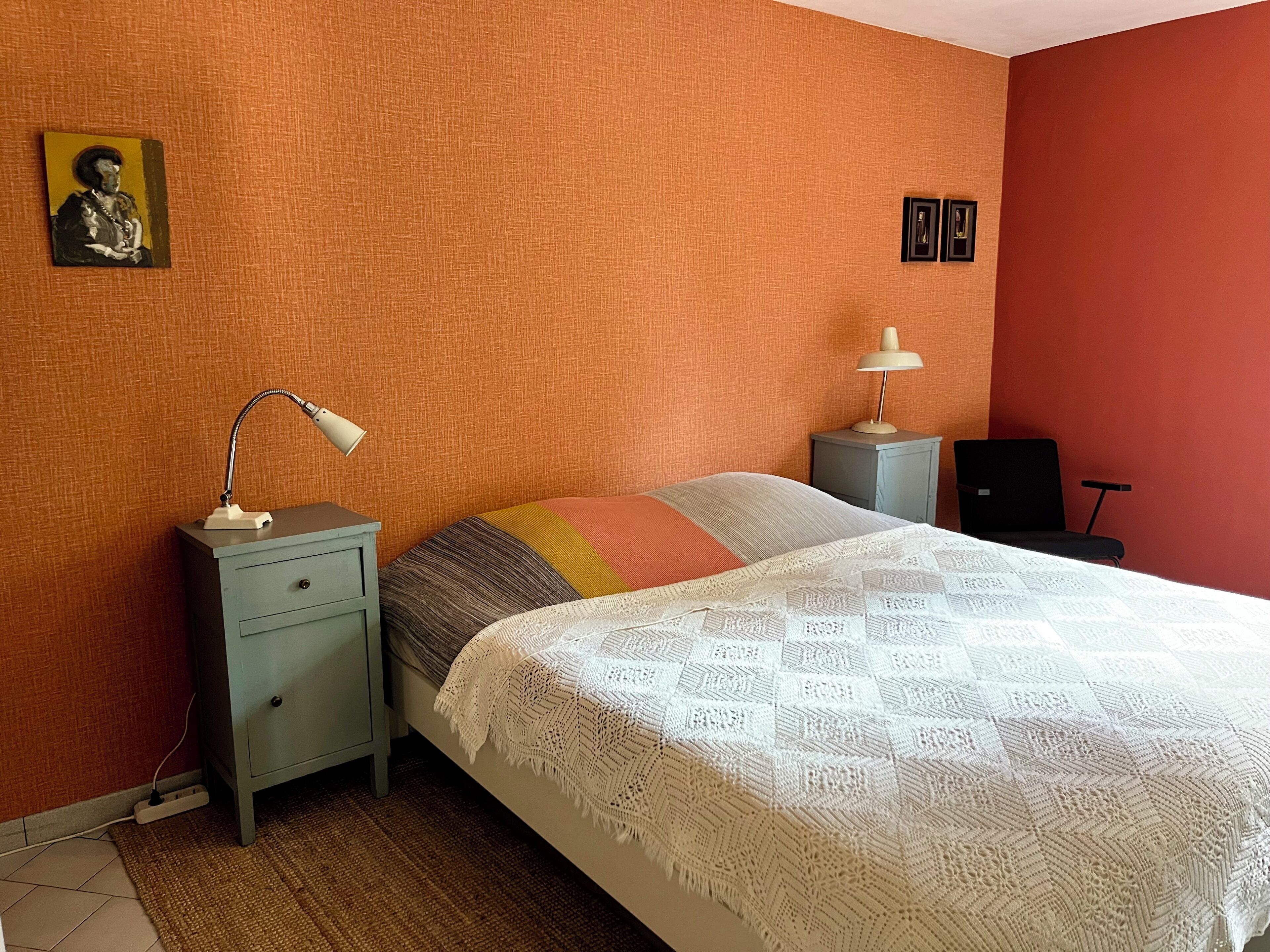 A bedroom with a bed, two bedside tables, a lamp, a power outlet, and a wall-mounted picture.
