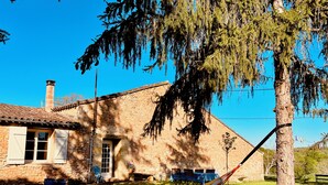 A rustic stone house with a tiled roof, a hammock, and a tree in the foreground. - Vue Épique : Gîte Magnifique (Capdrot)