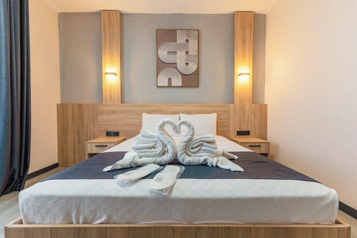 Een hotelkamer met een houten hoofdeinde, een bed met een witte matras en twee nachtkastjes met lampen.