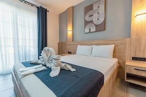 A neatly made bed with a blue and white bedspread, a wooden headboard, a wall-mounted abstract art piece, and a bedside table with a light switch. -  Nüans Küçükköy (Ayvalik)